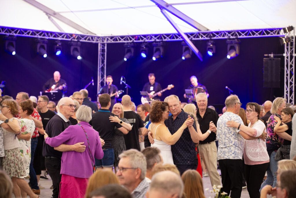 Tanssijat nauttivat livemusiikista suomalaisella tanssilavalla – parit tanssimassa ja orkesteri esiintymässä kesäillassa.