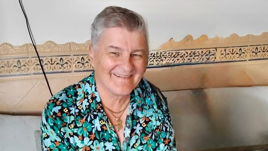 Hymyilevä mies kukkapaidassa istuu sisätilassa ja katsoo kameraan. Kuva: 2025.