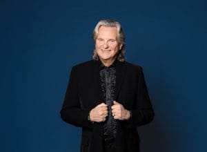 Pepe Willberg hymyilee kameralle mustassa juhla-asussa sinistä taustaa vasten. Kuvaaja: Satu Mali.