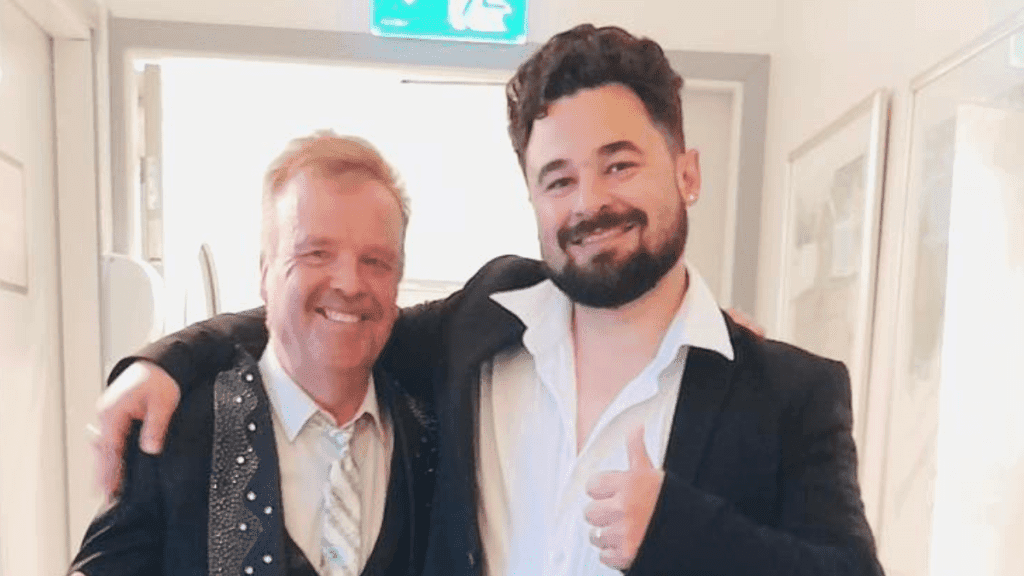 Two men in suits pose with arms around each other in a hallway; one gives a thumbs-up and both smile at the camera.