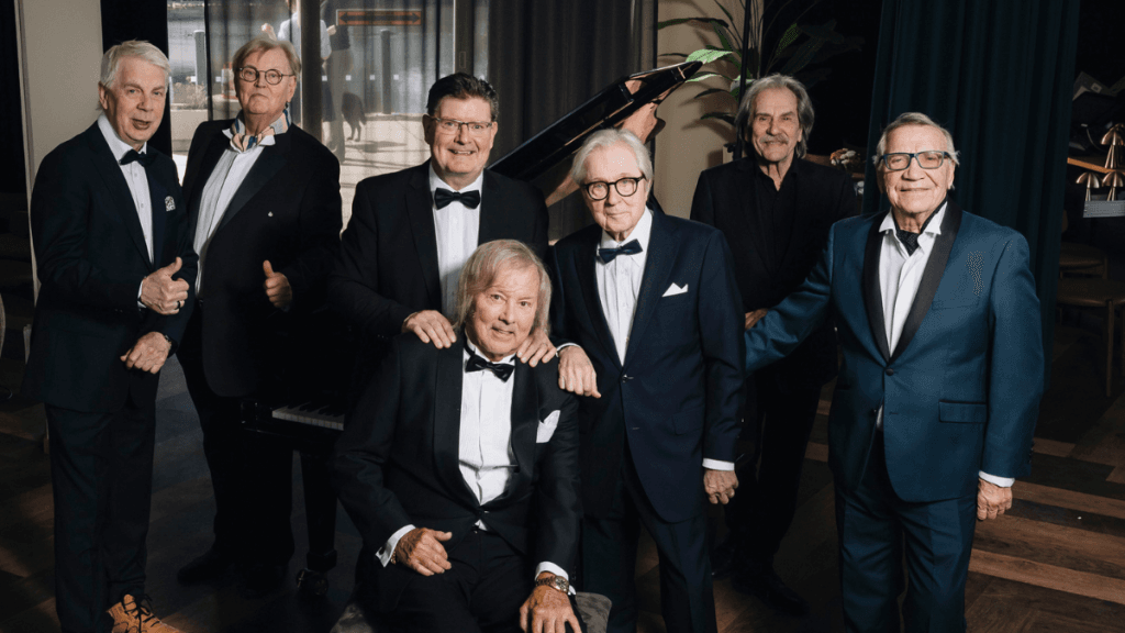 Eight men in tuxedos pose together around a grand piano in a stylish indoor venue, smiling for the camera.