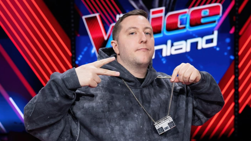 Man on a neon-lit stage wearing a dark hoodie, flashing a peace sign while holding a silver boombox pendant necklace.