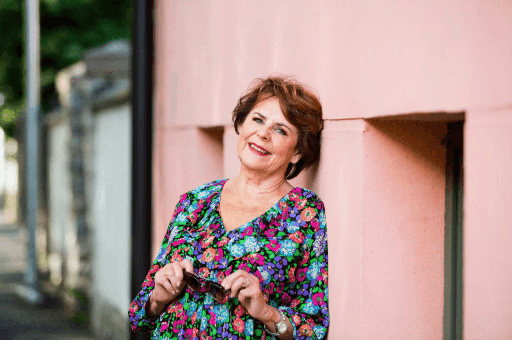 Smiling older woman leaning against a pink wall in a vibrant floral dress, holding sunglasses.