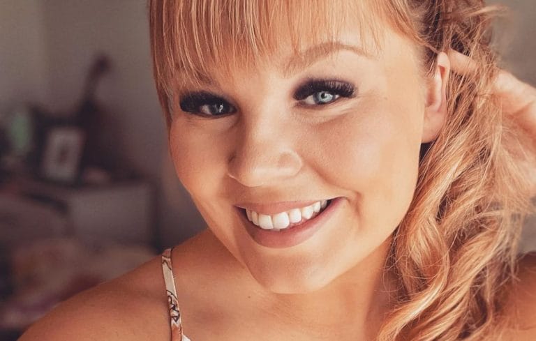 Close-up portrait of a smiling woman with blonde hair and blue eyes, wearing a floral-strap top.
