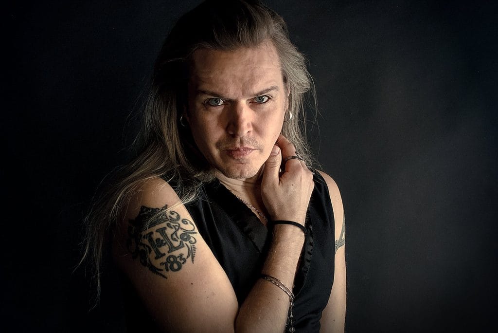Portrait of a man with long gray hair, wearing a black sleeveless shirt, looking at the camera with his hand near his chin; tattoos visible on his arm.