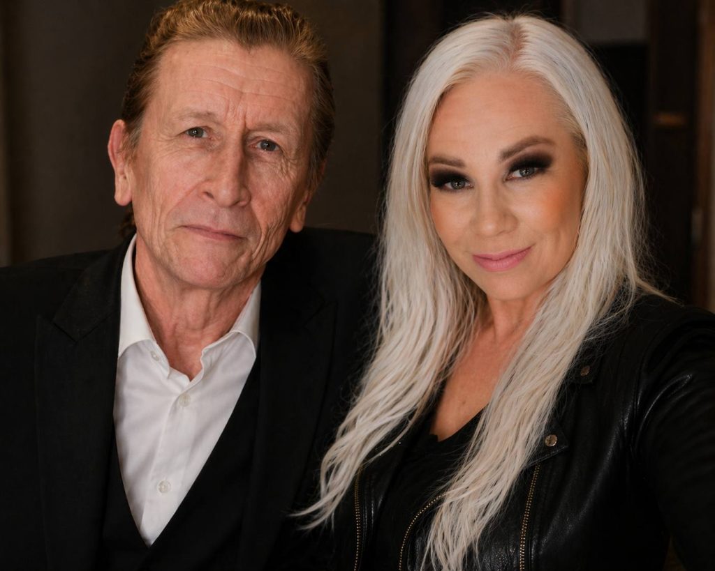 Two adults posing for a selfie: an older man in a white shirt and black blazer beside a platinum-blonde woman in a black leather jacket.