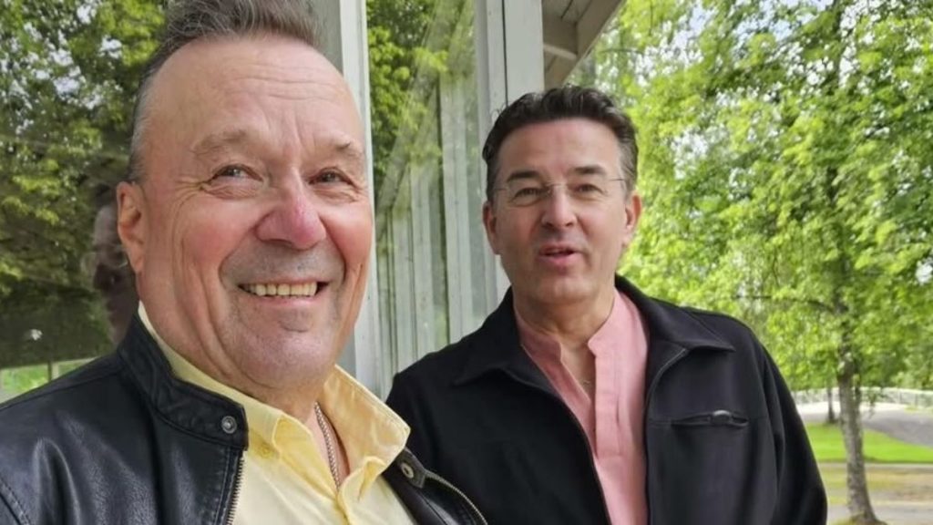 Two middle-aged men smiling at the camera outside, with a glass wall and green trees in the background.