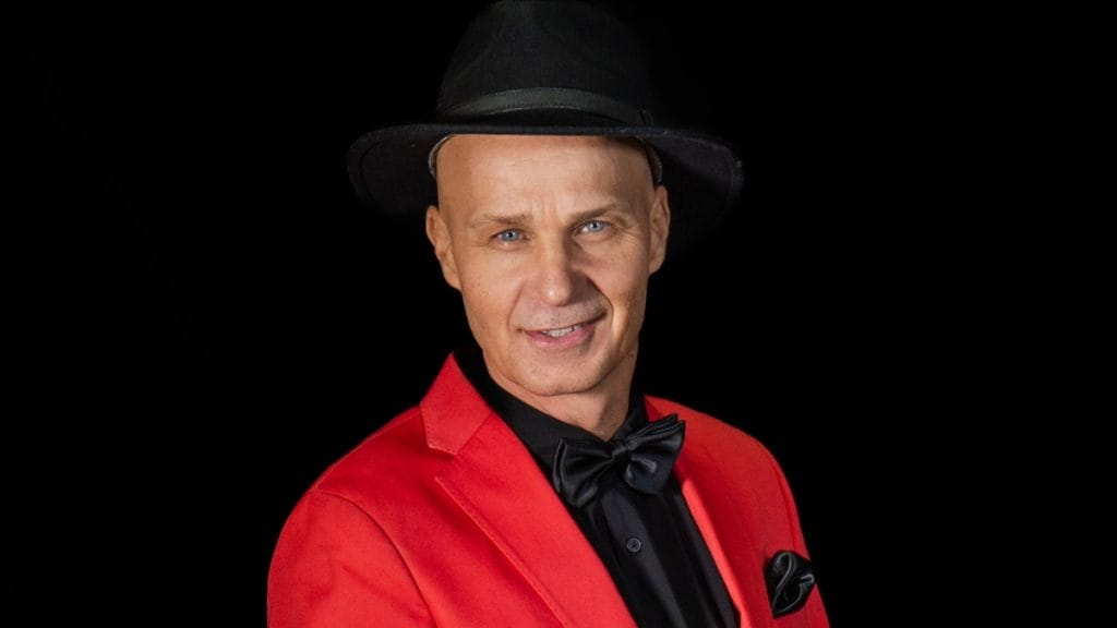 Smiling man in a red blazer, black shirt and bow tie, wearing a black hat against a dark background.
