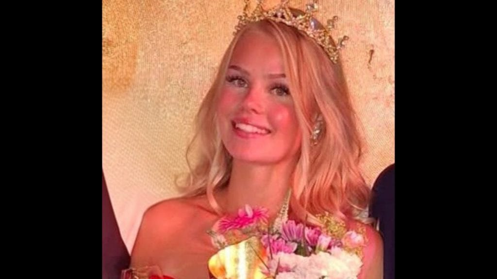 Smiling woman wearing a crown and holding a bouquet, likely a pageant winner, against a gold backdrop.