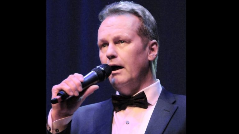 Male singer in a tuxedo belts into a microphone on stage.