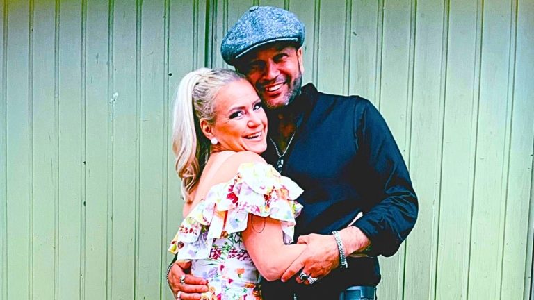 Smiling couple hugging outdoors in front of a green wooden wall; she wears a floral off-shoulder dress, he a flat cap and dark shirt.