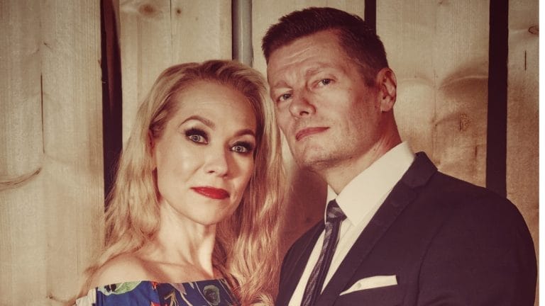 Couple posing for a photo: woman with blonde hair in a floral off-the-shoulder dress and red lipstick, man in a dark suit with white shirt and tie against a wooden background.