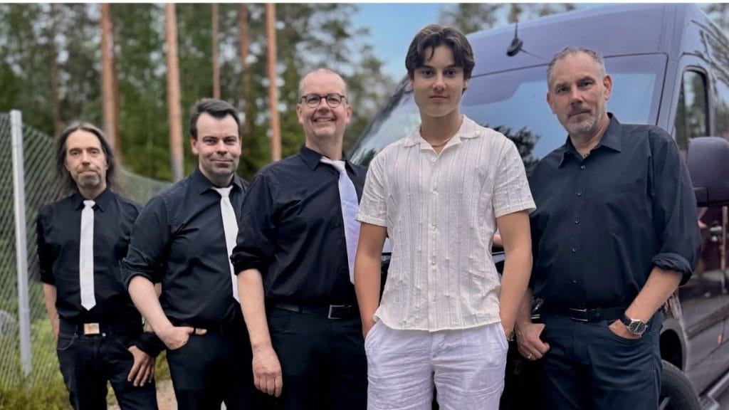 Five men pose in front of a dark van outdoors, wearing mostly black shirts; one younger man in a white linen short-sleeve shirt stands center-right.