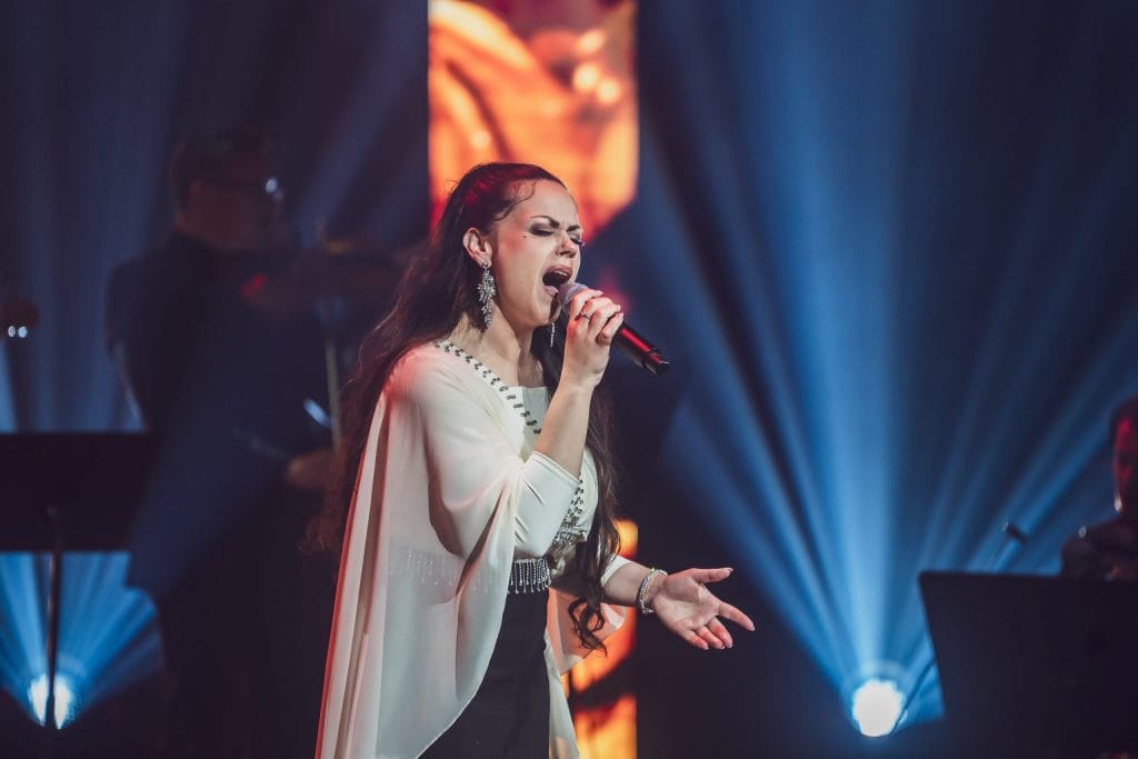 Female singer on stage wearing a white outfit, singing into a microphone with blue lighting in the background.