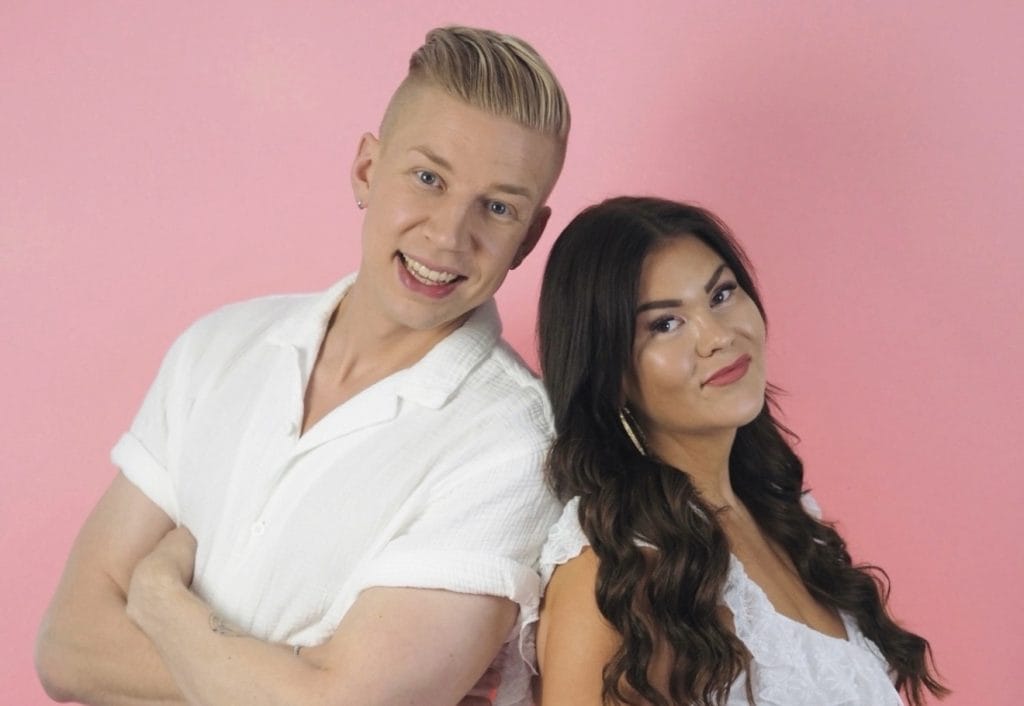 Two people pose back-to-back against a pink backdrop; man wearing a white shirt and woman in a white dress, both smiling at the camera.