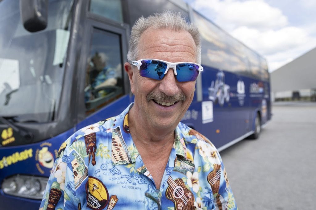 Smiling man in a Hawaiian shirt with blue mirrored sunglasses taking a selfie in front of a blue tour bus.
