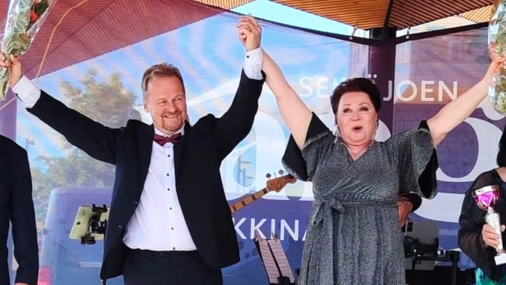 Man and woman on a stage, arms raised in celebration, each holding a bouquet, with a guitarist and banner in the background.