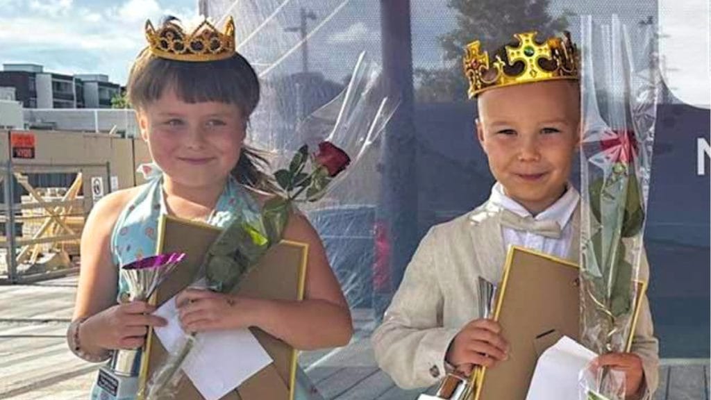 Two young children wearing crowns stand with flowers and certificates outdoors, celebrating a graduation or pageant.