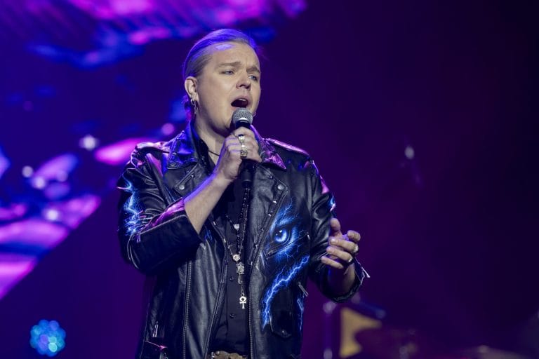 Male singer performing on stage in a black leather jacket, microphone in hand, illuminated by purple concert lighting.