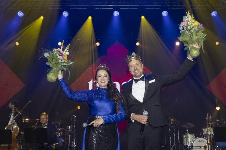 Man and woman in formal attire wearing crowns celebrate on stage, each holding a bouquet raised in the air.
