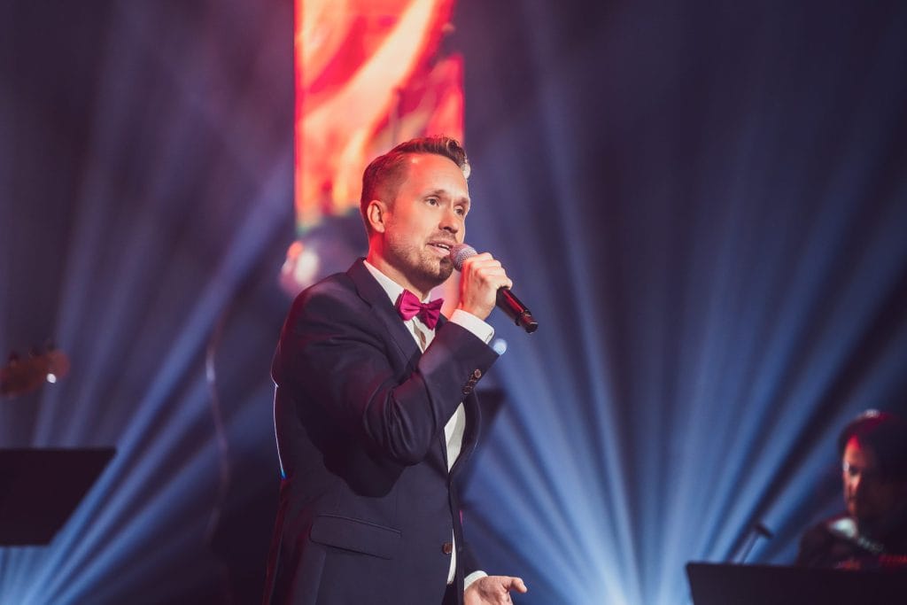 Male singer in a dark suit and magenta bow tie performs on stage, holding a microphone with dramatic blue spotlighting in the background.