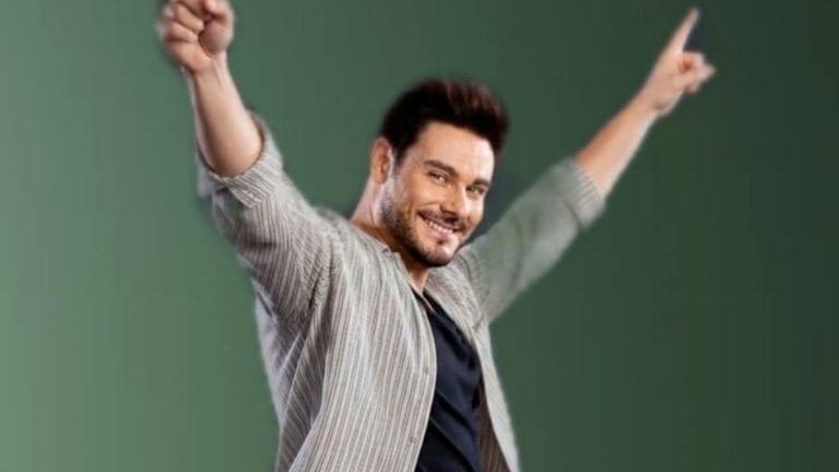 Man in a striped shirt raises both arms in celebration, smiling at the camera against a green background.