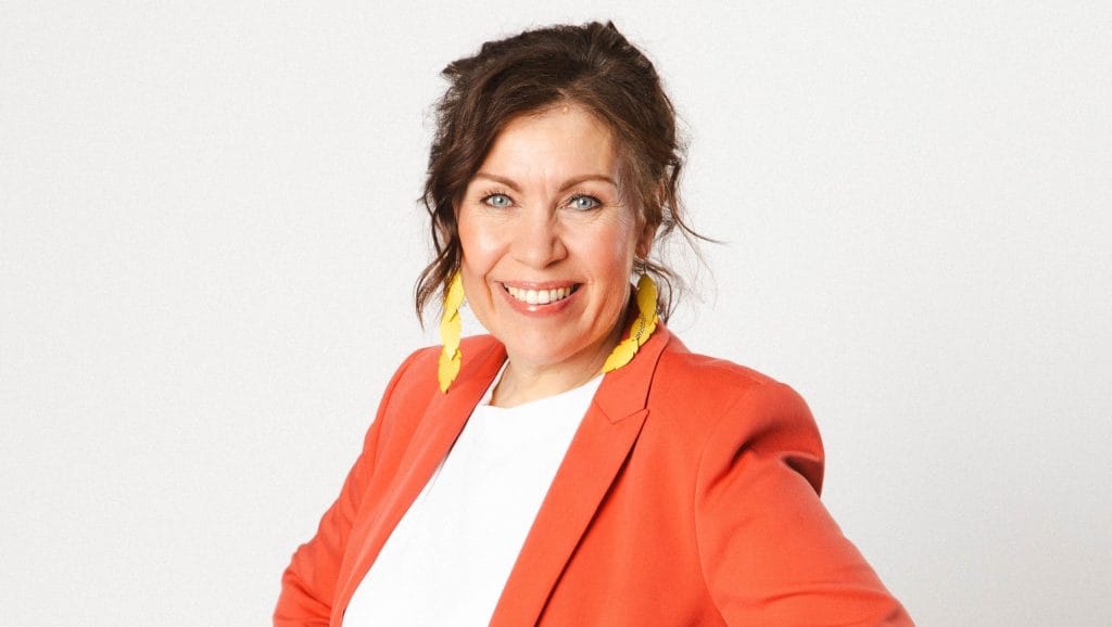 Smiling woman in an orange blazer with yellow earrings, facing the camera against a light background.