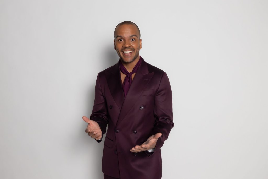 Smiling man in a burgundy suit with open hands against a light gray background
