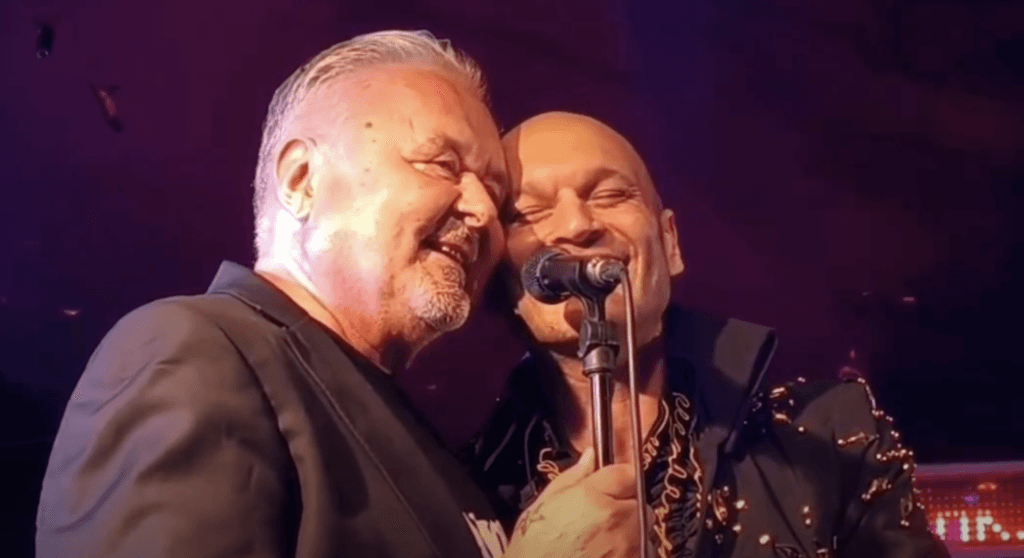 Two male singers sharing a microphone on stage, smiling and leaning close together as they perform.