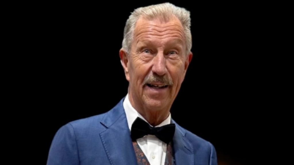 Older man with gray hair and mustache, wearing a blue suit and black bow tie, smiling against a black background.