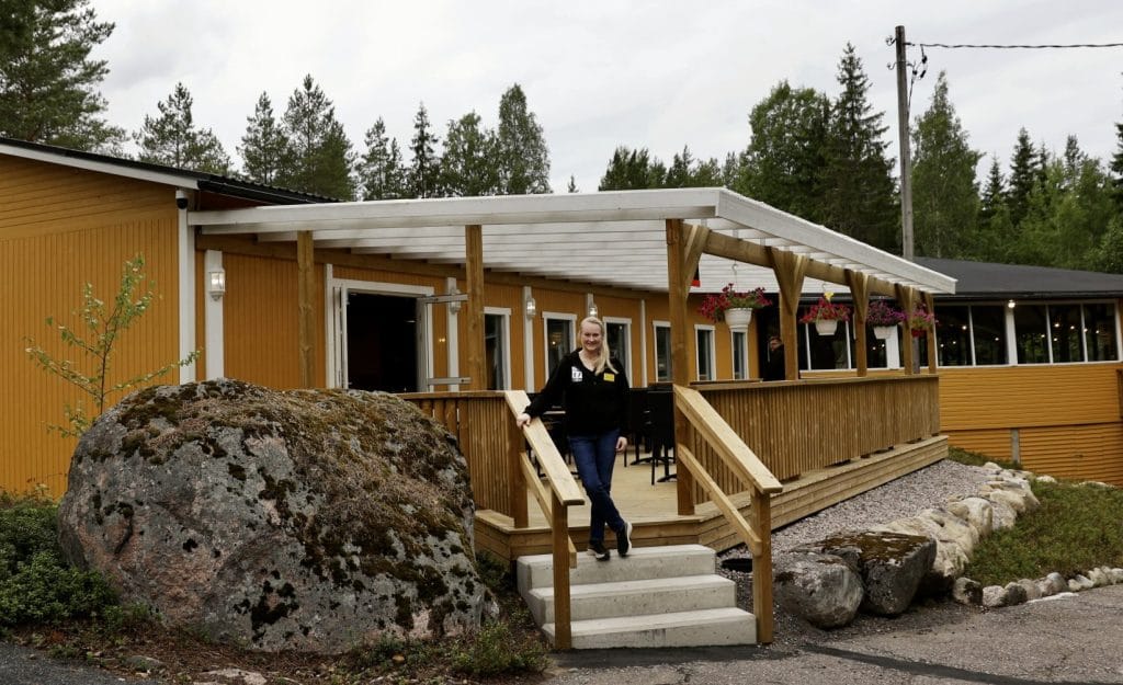 Yellow wooden building with a purple sign and people on the covered porch; a woman stands near the railing by a large rock in front.