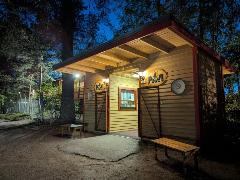 Small wooden kiosk with yellow circular signs and warm lights at night, surrounded by trees and a chain-link fence; benches outside.