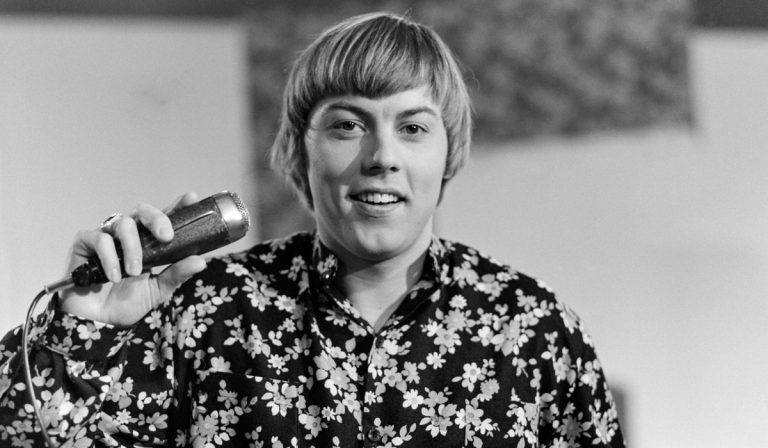 Person with short hair wearing a floral shirt speaks into a handheld microphone.