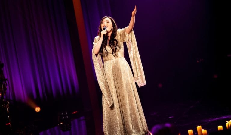 Female singer on a stage in a gold shimmering gown, mic in hand, one arm raised during a performance with purple stage lighting.