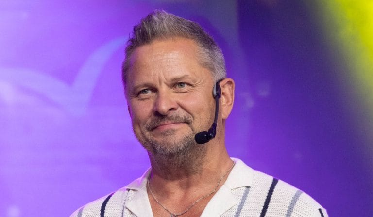 Male presenter on stage wearing a headset microphone, smiling at the audience with purple stage lighting behind him.