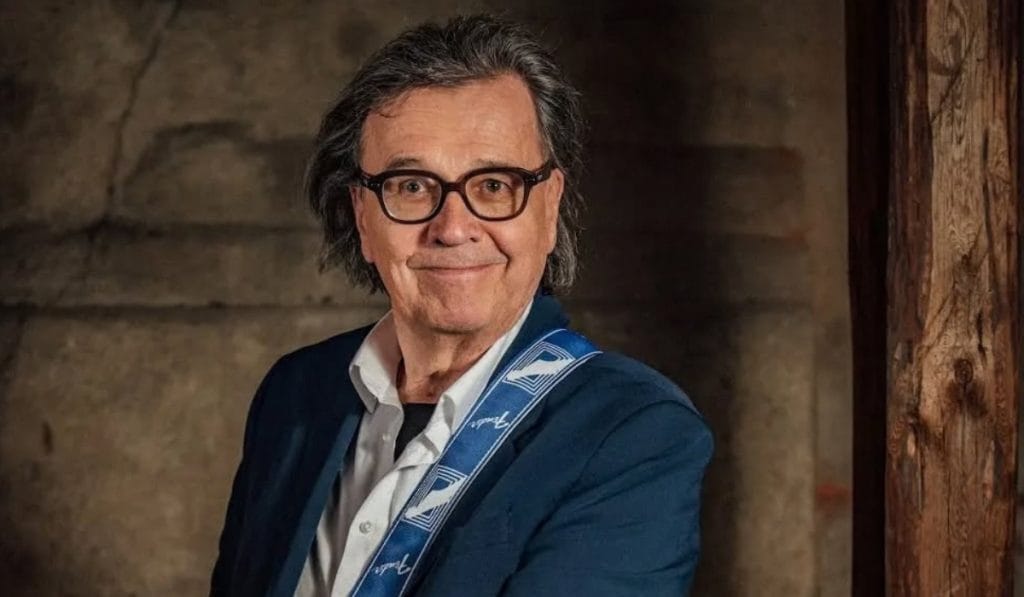 Smiling man with dark hair and glasses wearing a blue blazer and white shirt, a blue guitar strap over his shoulder.